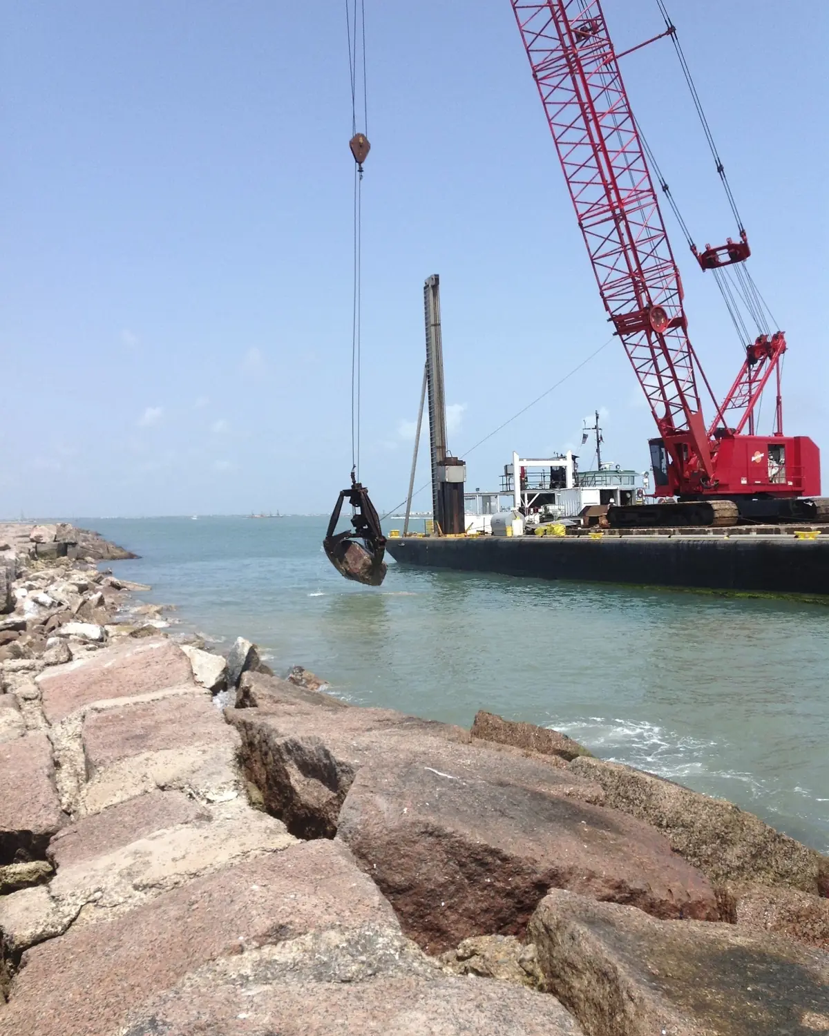 Red crawler crane on a barge working on the rock jetty at the entrance to the Brownsville Ship Channel