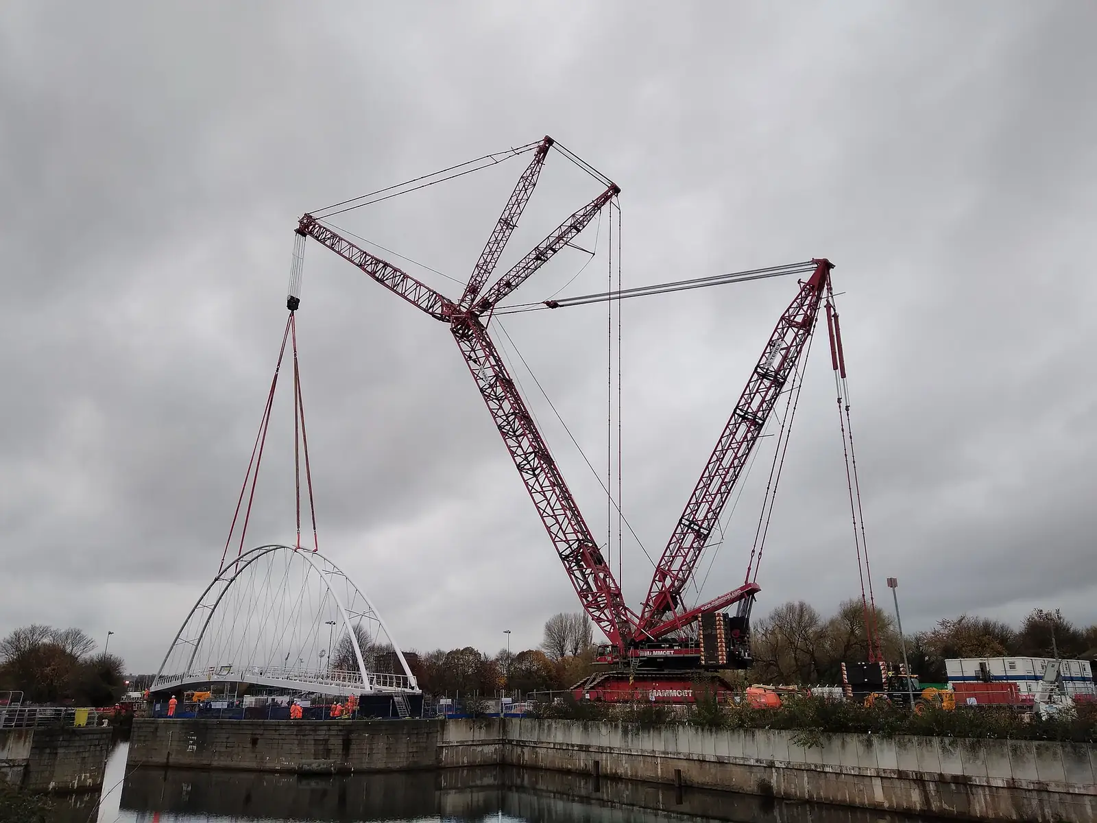 Large red lattice-boom crawler crane lifting a white steel arch footbridge over a river