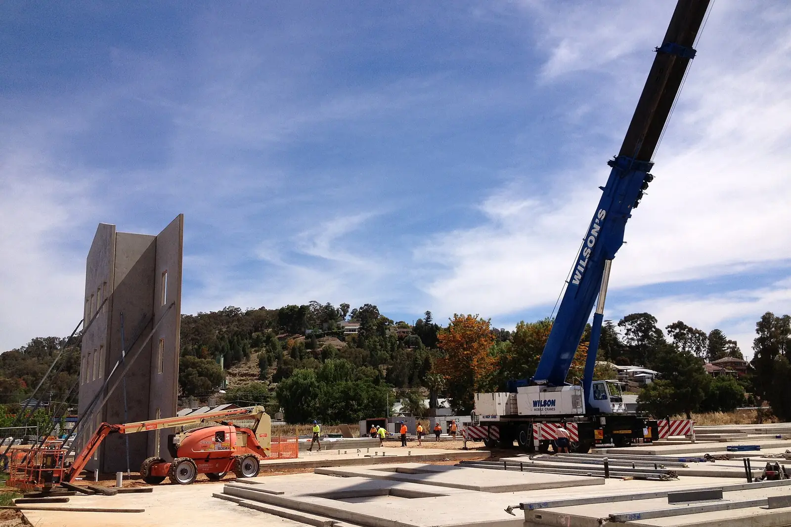 Blue mobile crane on outriggers lifting a tall precast concrete wall panel on a building site