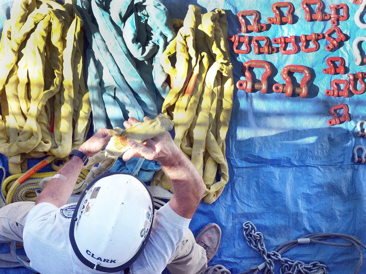 A rigger in a hard hat sorting yellow round slings and red shackles laid out on a blue tarp