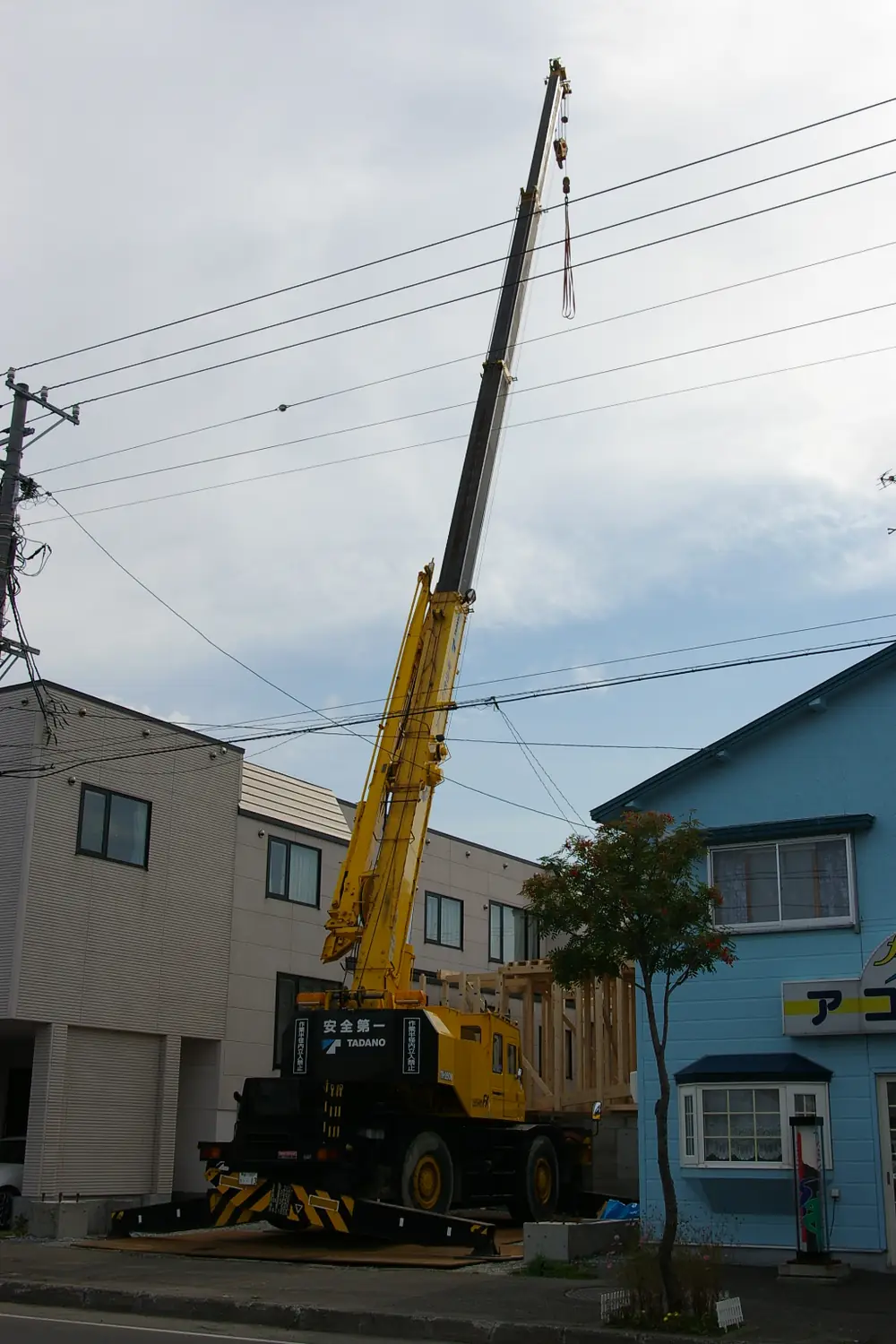 Yellow rough-terrain crane with its boom raised high, set up on timber mats between two buildings beside a street